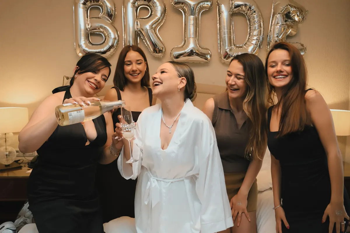 The bride in a white robe toasting under BRIDE letter balloons